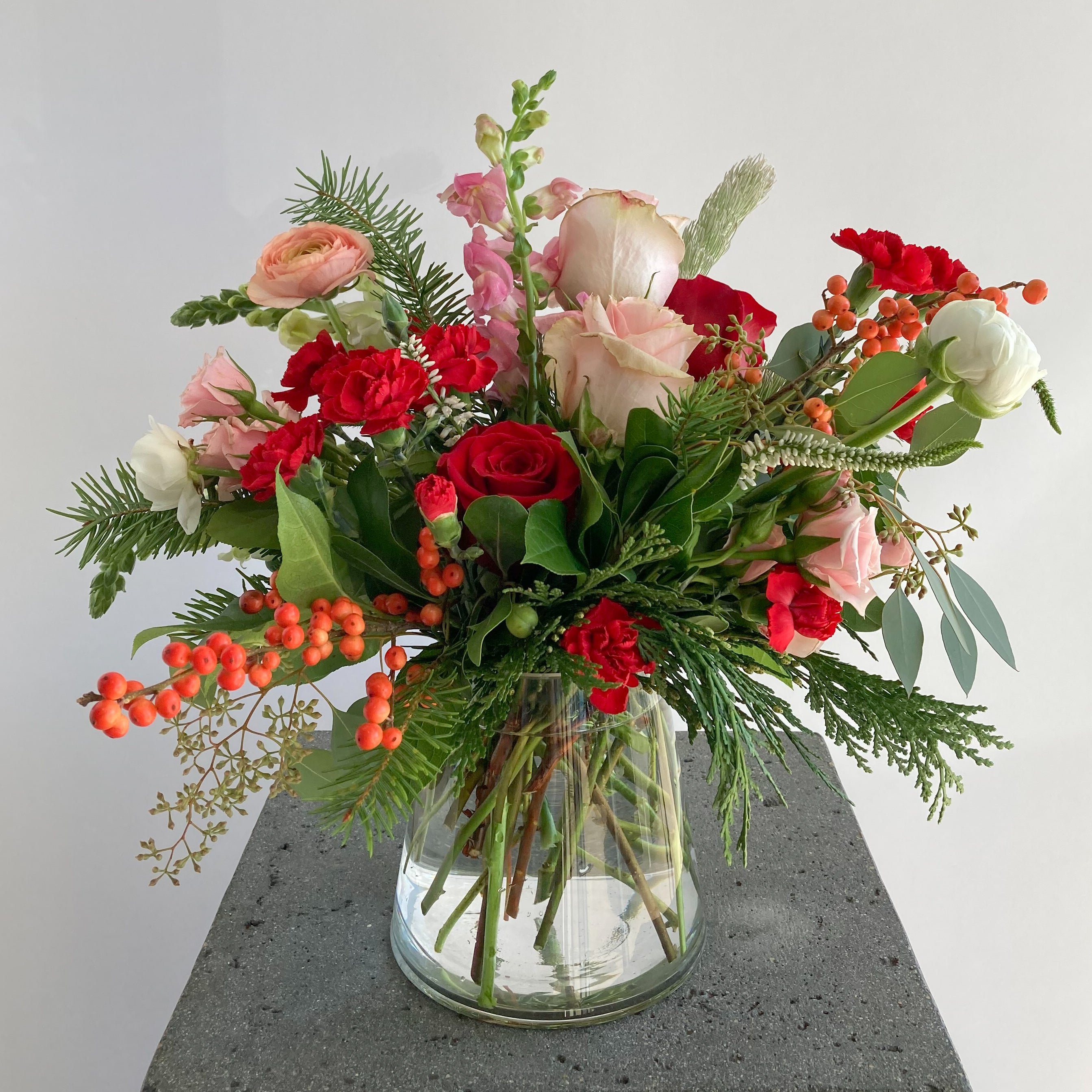 Bouquet of red, pink, and white flowers with Christmas greens in a clear vase 