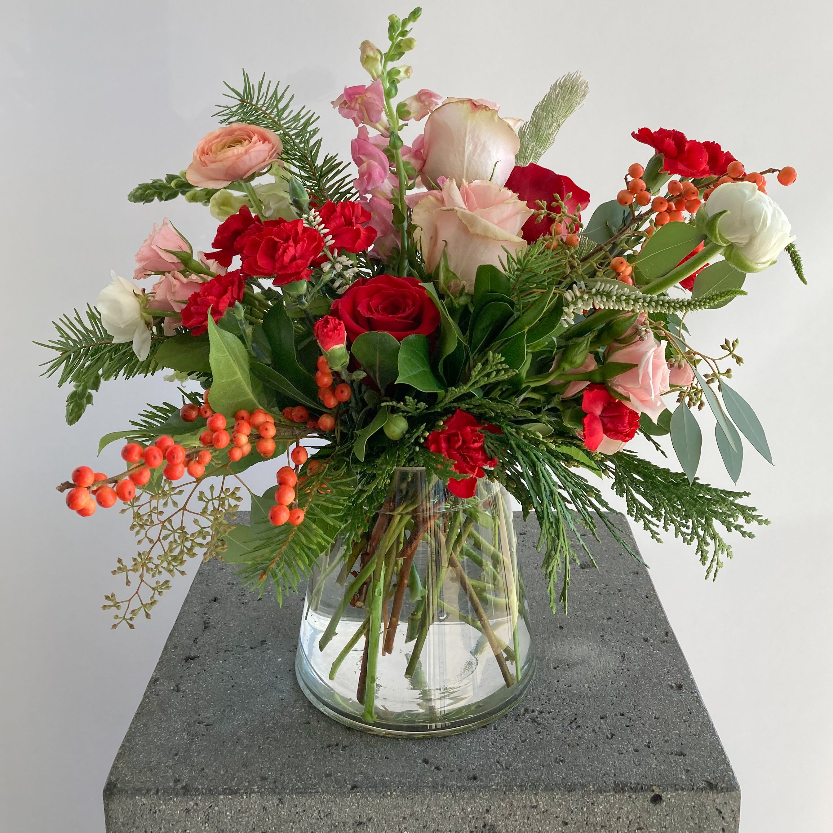 Bouquet of red, pink, and white flowers with Christmas greens in a clear vase
