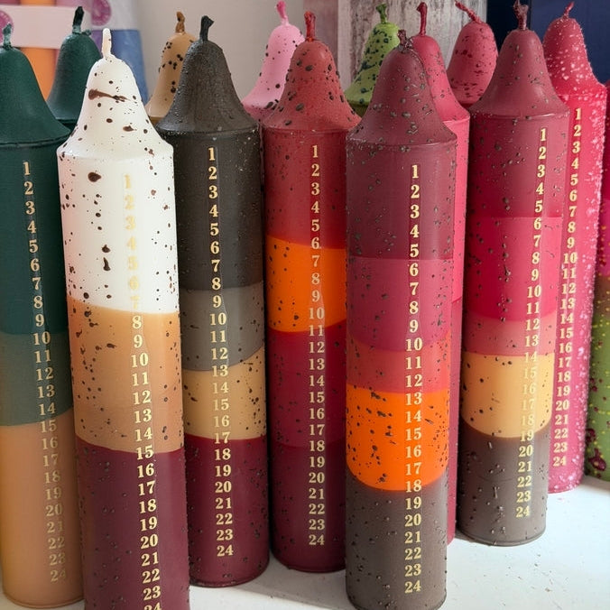 A set of colorful taper Advent candles arranged upright, each printed with numbered days from 1 to 24. The candles feature various color-blocked patterns in pink, red, orange, brown, beige, and green, with small black speckles.
