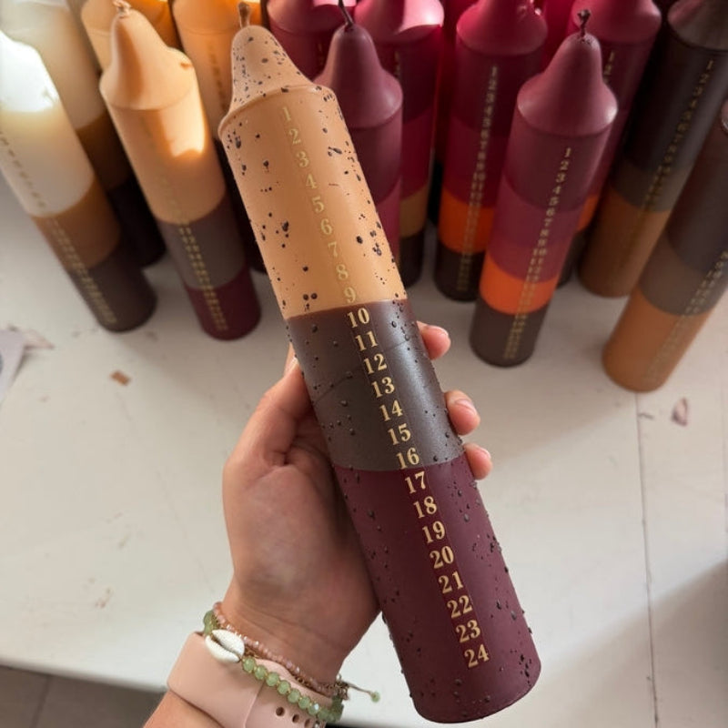 A hand holds a 24-day advent candle, which is speckled beige at the top and dark brown at the bottom with gold numbers. 