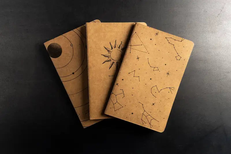 Three brown notebooks with star and constellation designs on a dark surface