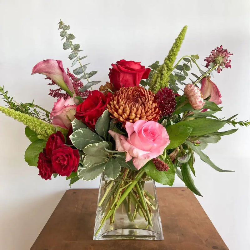 Bouquet of red, pink, and green flowers in a clear vase on a wooden surface with a white background