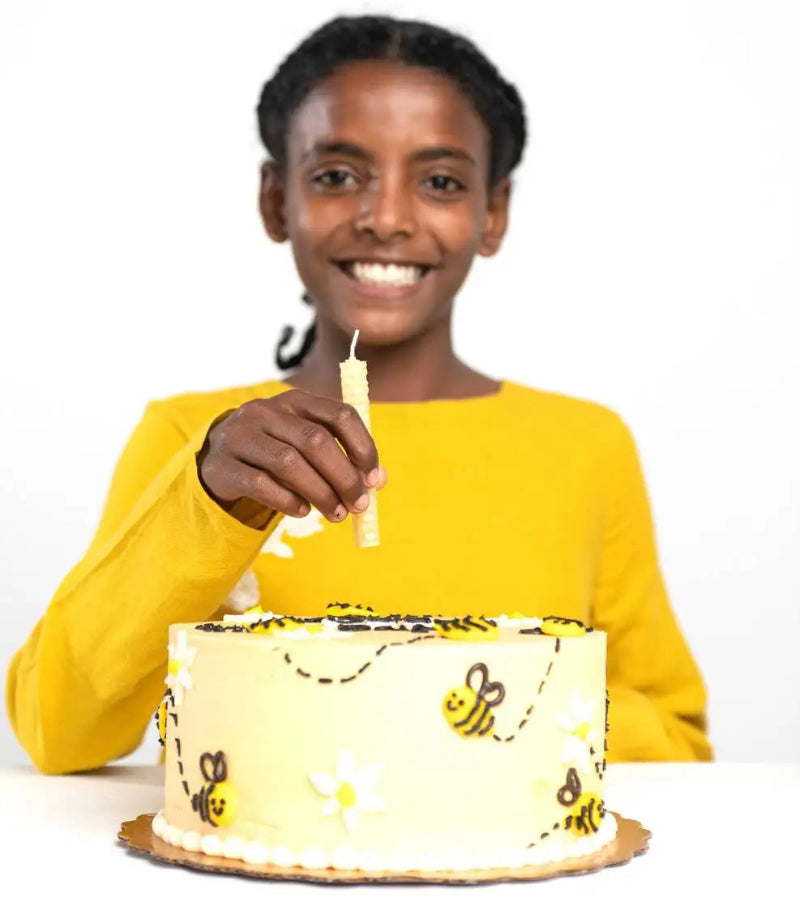 A smiling woman in a yellow long-sleeve shirt holds a single lit candle above a round birthday cake decorated with yellow and black bumblebees and white flowers on a white background.