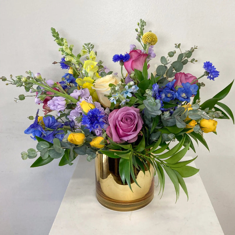Colorful flower arrangement in a gold vase on a white surface with a light gray background
