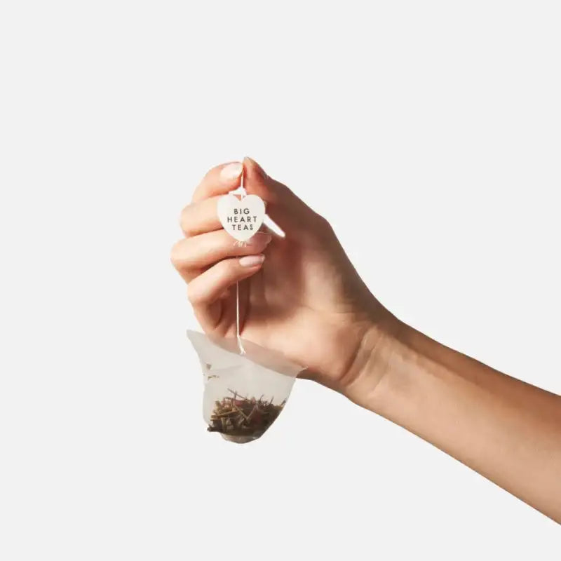 Hand holding a big heart tea bag with visible tea leaves against a white background