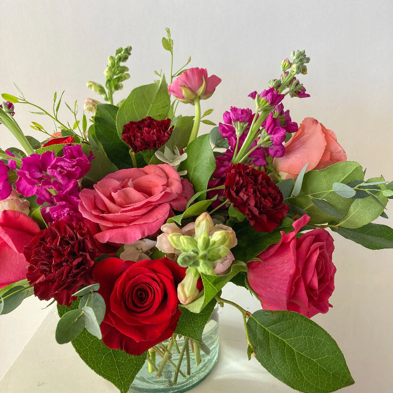Bouquet of colorful flowers including red, pink, and purple roses with green leaves in a clear vase on a light background.