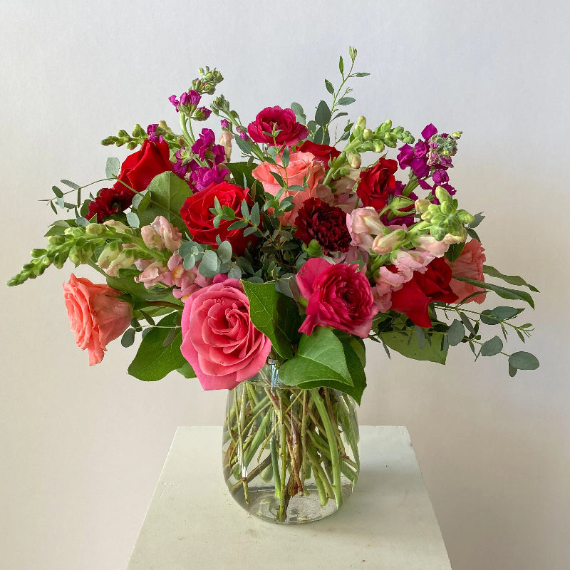 Bouquet of red, pink, and green flowers in a clear vase on a white surface with a light gray background