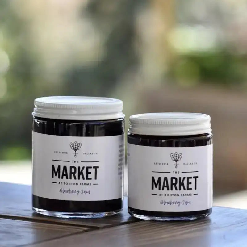 Two jars of 'Blueberry IPA' from 'The Market at Bonton Farms' on a wooden surface with a blurred natural background.