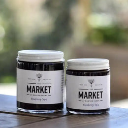 Two jars of 'Blueberry IPA' from 'The Market at Bonton Farms' on a wooden surface with a blurred natural background.
