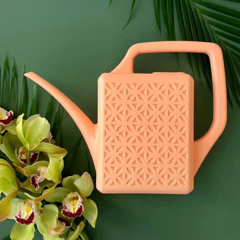 Peach-colored watering can with a geometric pattern on a green background with orchids.