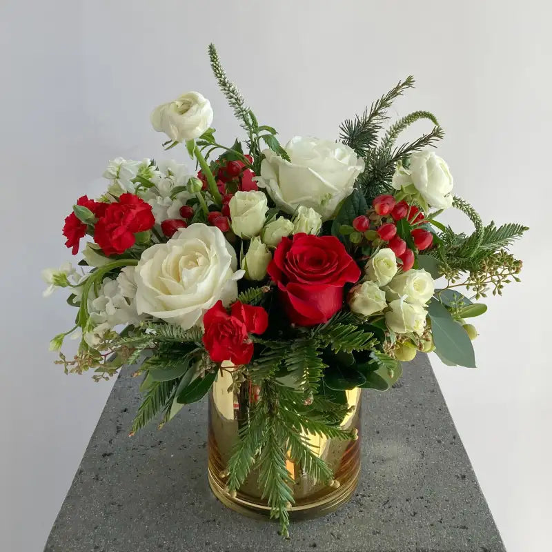 Bouquet of red, white, and green flowers in a gold vase on a gray surface with a light gray background.