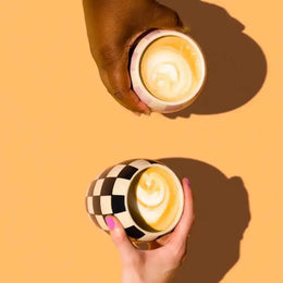 Two hands holding checkered coffee cups with latte art against a yellow background
