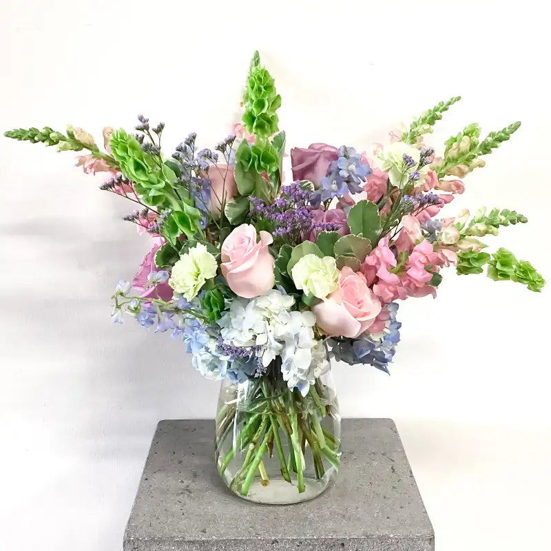 A floral arrangement featuring a variety of flowers including roses and other greenery, in pastel colors, placed in a tall clear vase.