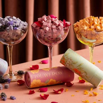 Three glass bowls filled with colorful flower-infused cocktail cubes on a pink surface with 'FlowerCo' branding.