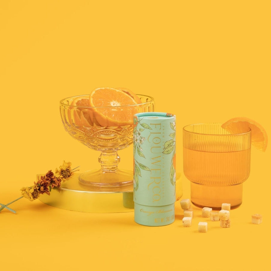 A styled product shot on a solid yellow background featuring a mint green cylindrical container of floral-infused cocktail cubes, a clear glass bowl with orange slices, a textured orange-tinted glass, and scattered cocktail cubes.
