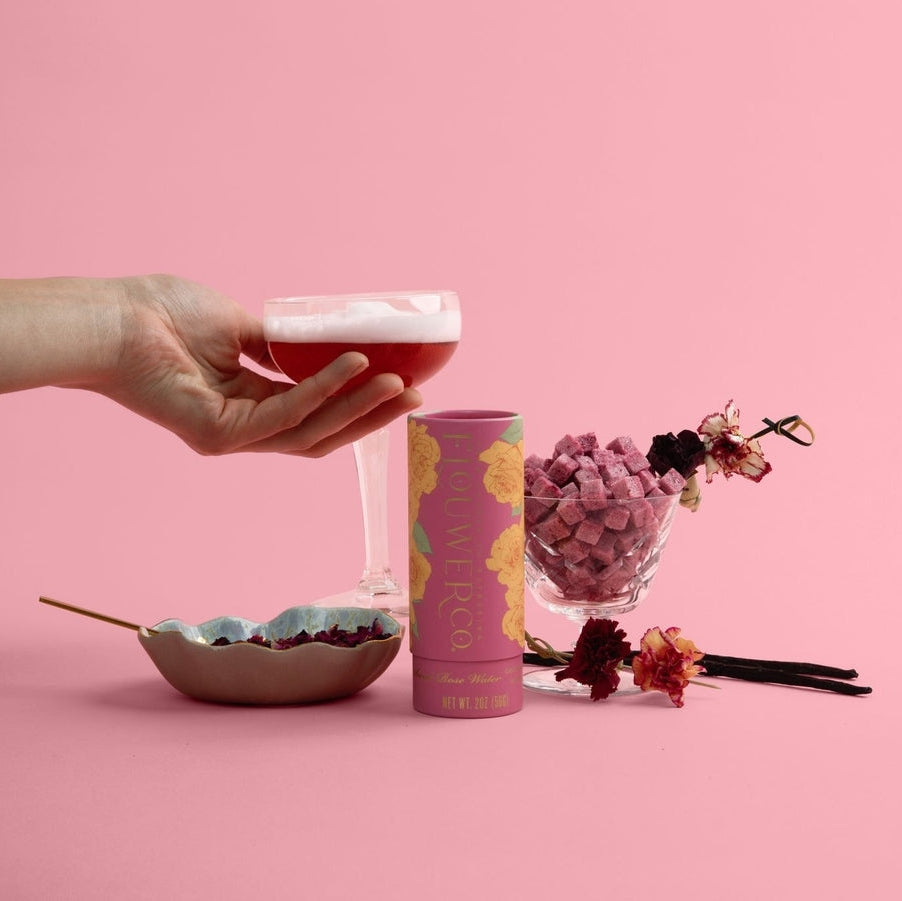 Hand holding a glass of pink champagne with a pink background, featuring a bowl, floral-infused cocktail cubes, and flowers.