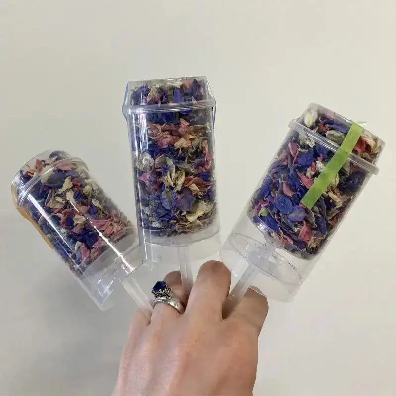 Three clear containers filled with colorful dried flowers, held by a hand against a white background.