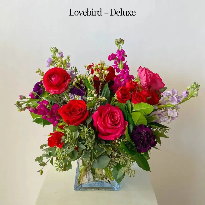Bouquet of red, pink, and purple flowers in a clear vase on a light background with 'Lovebird - Deluxe' text.