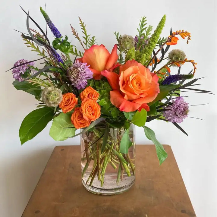 Bouquet of orange roses and other flowers in a clear vase on a wooden surface.