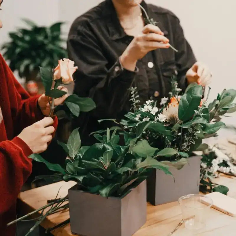 Two people arranging flowers in gray containers on a wooden table.