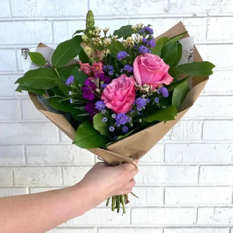 Handheld bouquet of pink roses with purple asters and greenery wrapped in brown craft paper against a white brick background
