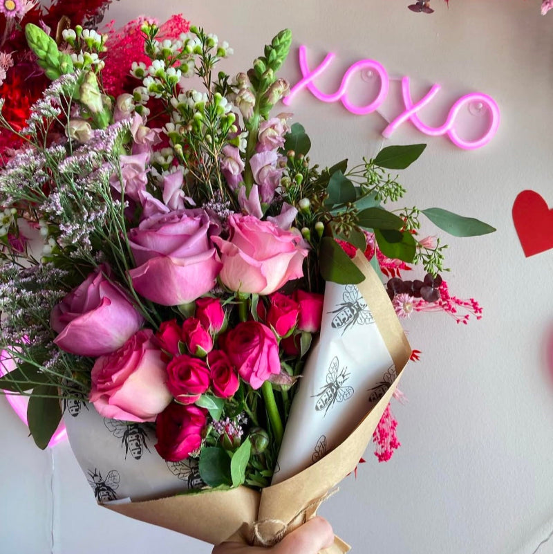 A wrapped bouquet of pink and red flowers with greenery, presented in front of a pink neon sign and various decorative elements.
