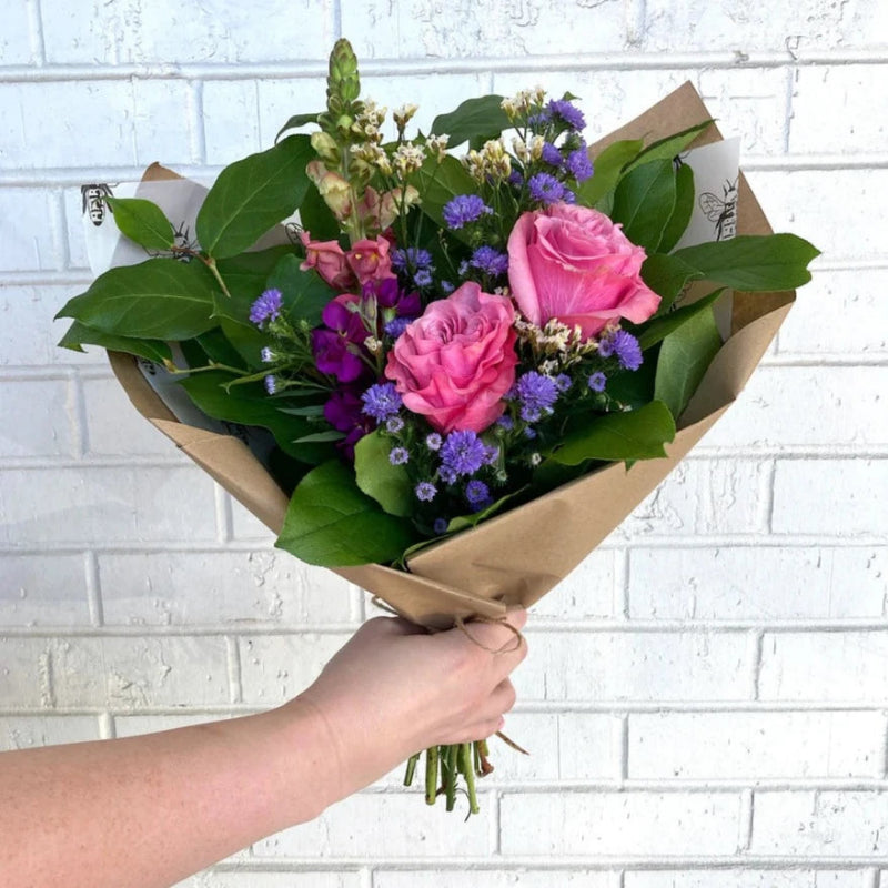 Handheld bouquet of pink roses with purple asters and greenery wrapped in brown craft paper against a white brick background