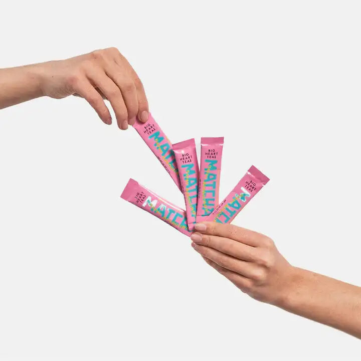 Two hands holding pink packets with 'Matcha' text on a light gray background