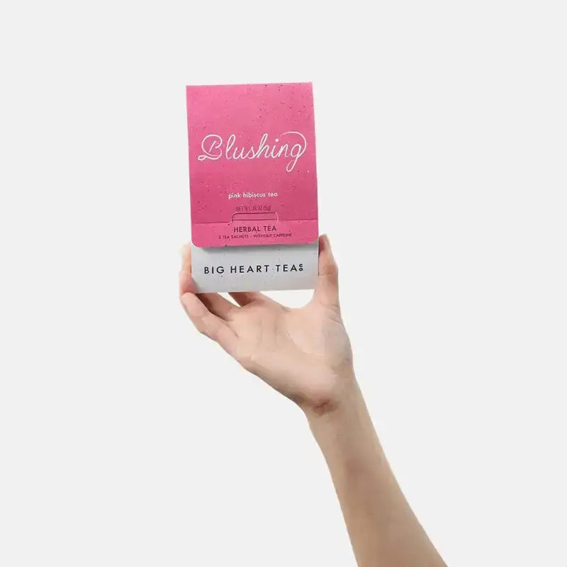 Hand holding a pink package of 'Blushing' herbal tea from Big Heart Tea on a white background