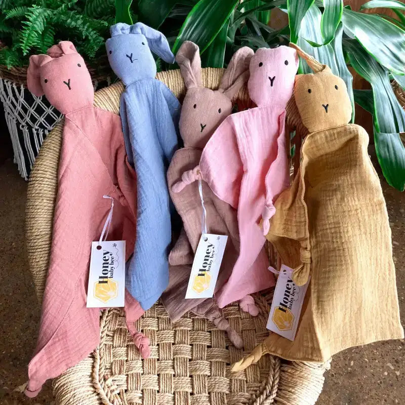 Five soft toy rabbits in different colors with tags on a woven surface.