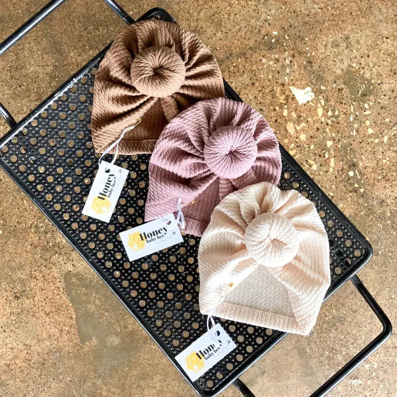 Three velvet baby hats in brown, pink, and beige on a black textured surface with 'Honey' brand tags.