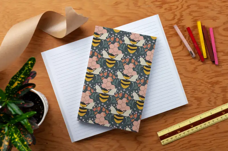 Notebook with floral and bee pattern on a wooden surface with stationery items.