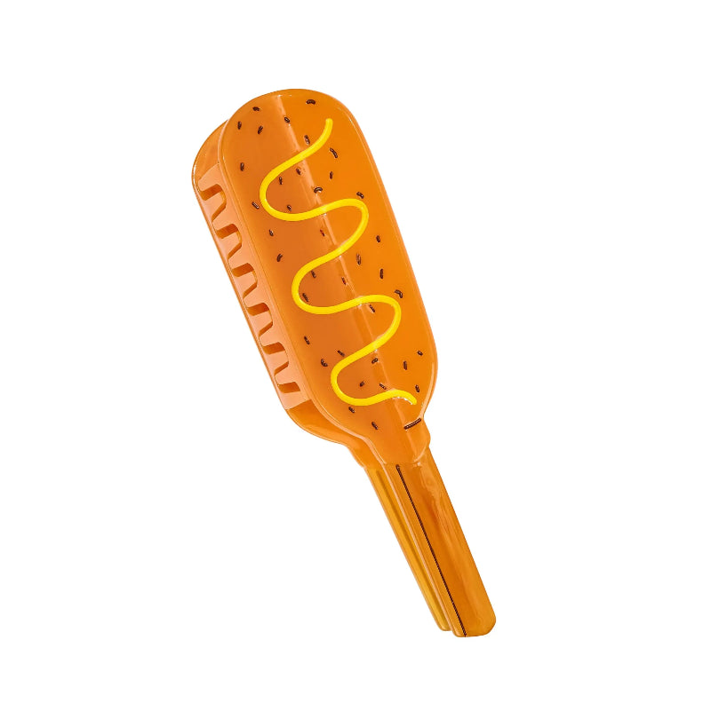 An orange plastic toy brush shaped like a hot dog bun with a yellow wavy line representing a hot dog on top, isolated on a white background.
