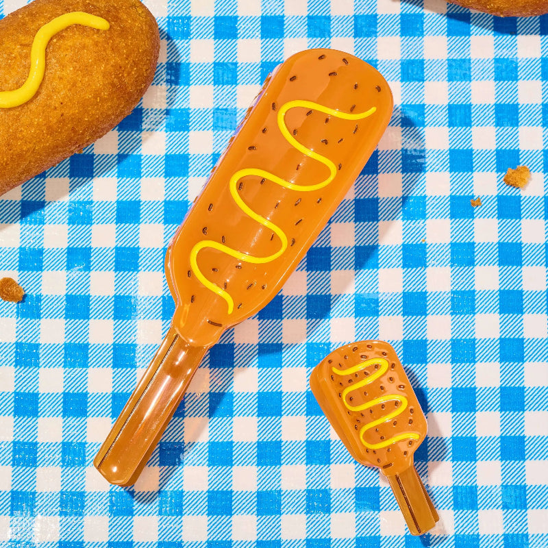 Two translucent orange corn dog-shaped utensils or tools with yellow mustard-like wavy detailing and small black speckles, placed on a blue-and-white checkered surface with partial fried food props visible.