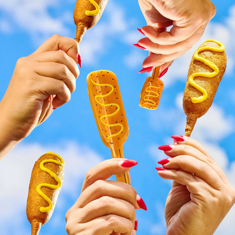 Multiple hands with red manicured nails hold various golden-brown corn dogs and a yellow mustard-covered skewer against a bright blue sky with white clouds.