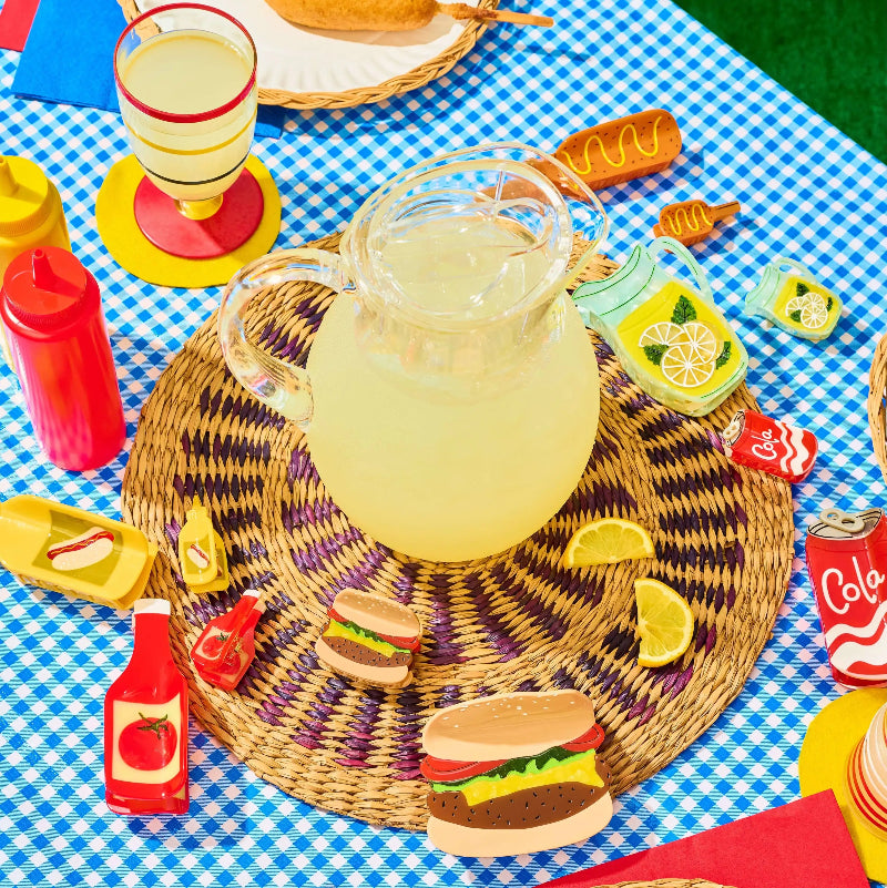 A styled picnic tabletop scene with a clear glass pitcher filled with pale yellow drink on a woven round placemat, surrounded by colorful toy condiment bottles, burgers, lemon slices, napkins, and a blue-and-white checkered blanket.
