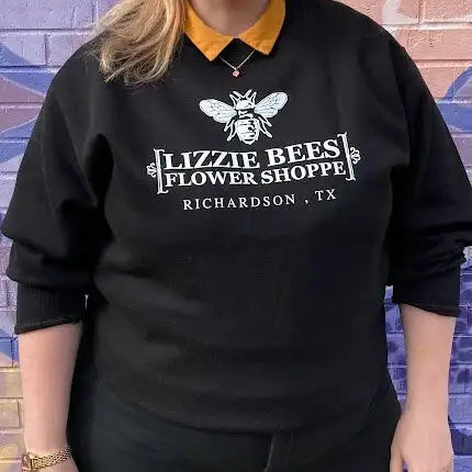 Person wearing a black sweatshirt with 'Lizzie Bees Flower Shoppe' logo against a colorful brick wall.