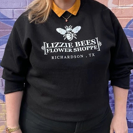 Person wearing a black sweatshirt with 'Lizzie Bees Flower Shoppe' logo against a brick wall.