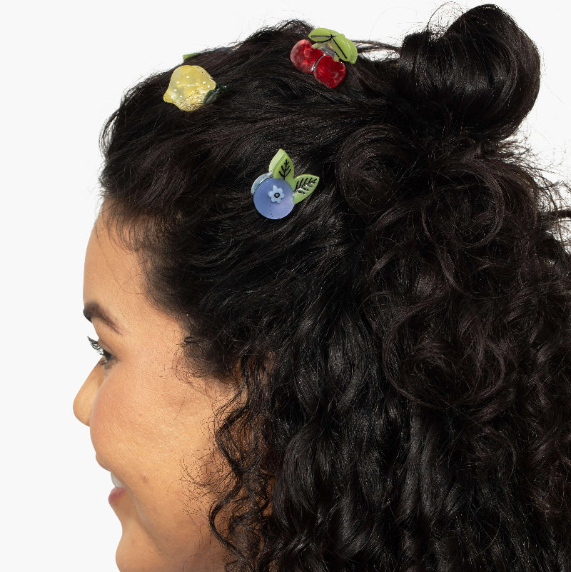 A close-up side profile of a person with dark curly hair wearing three colorful hair clips shaped like a lemon, cherries, and a blueberry, set against a plain white background.