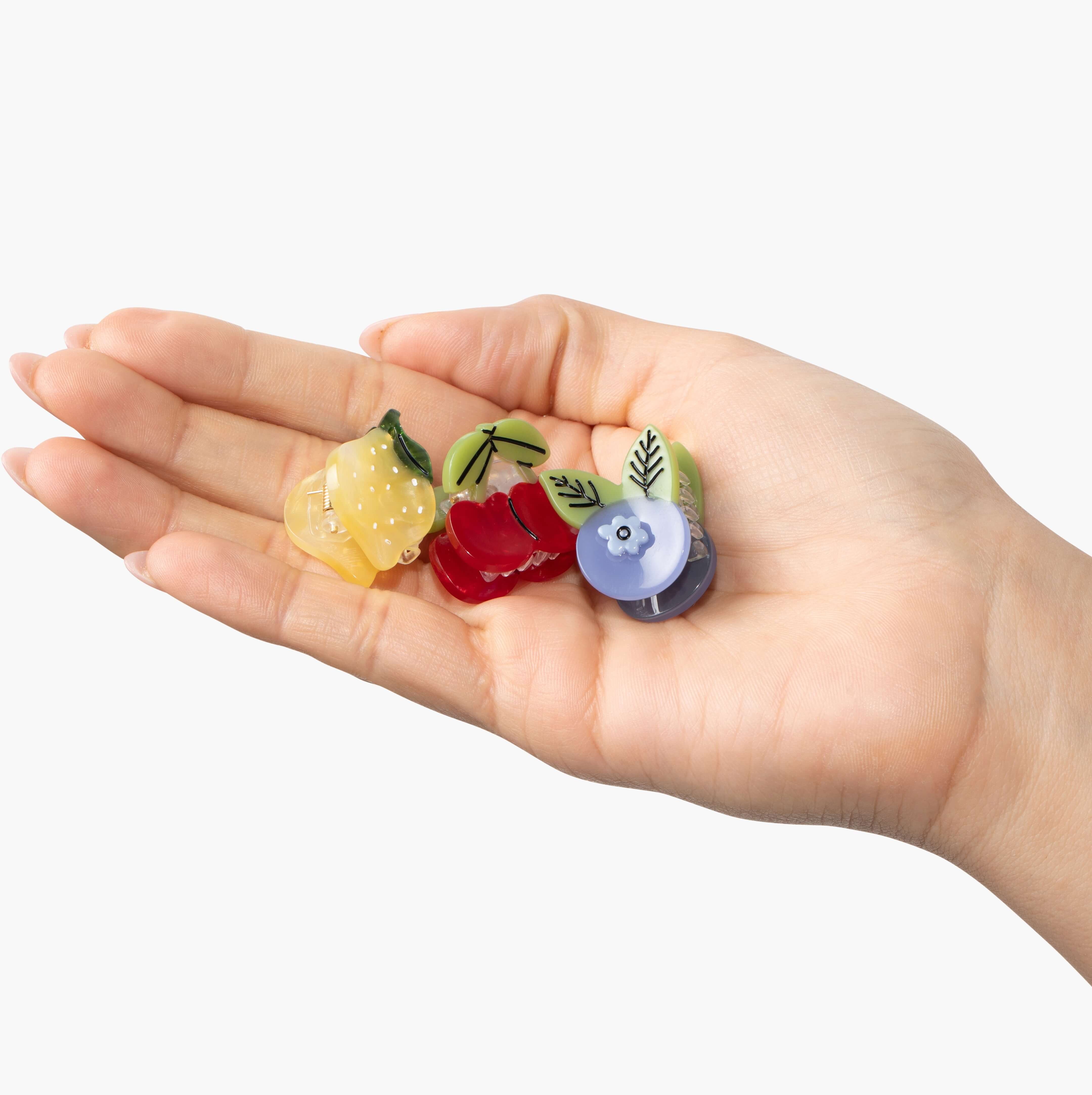 Three small claw hair clips held in an open palm. One lemon, one cherry, one blueberry hair clip.