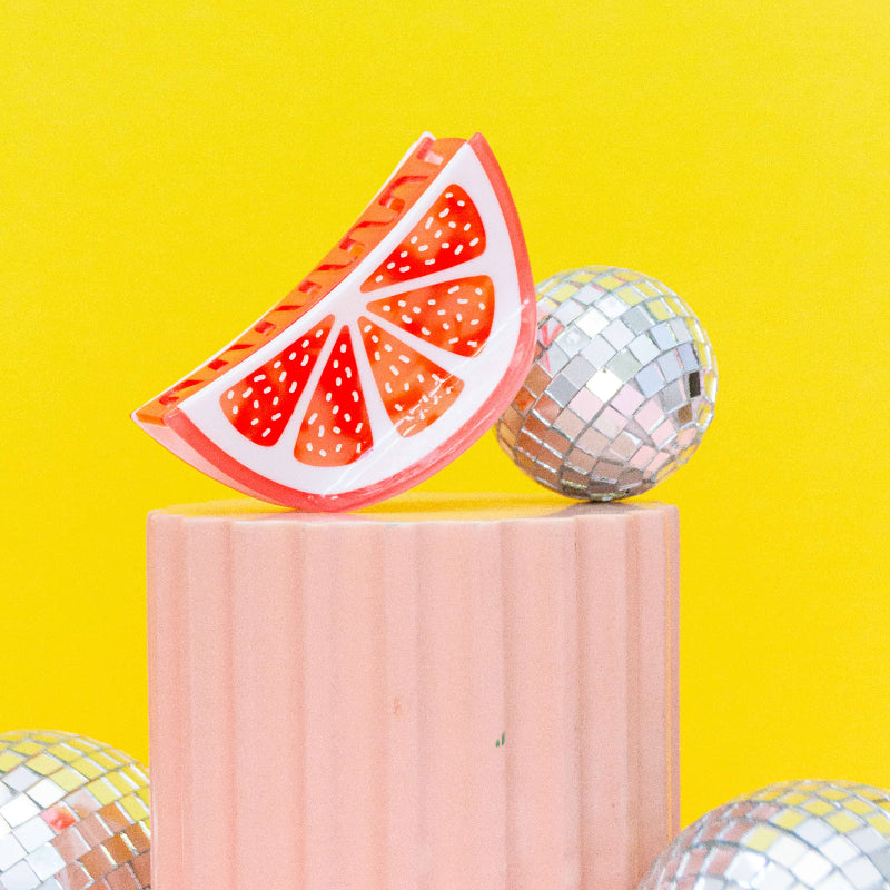 Grapefruit slice hair claw clip and disco ball on a pink pedestal against a yellow background