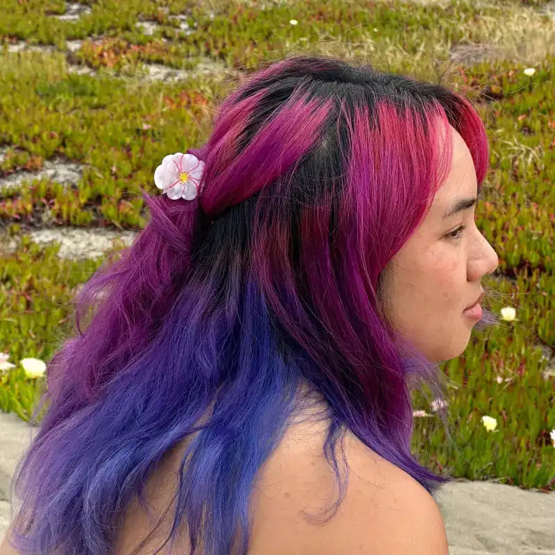 Person with vibrant purple and pink hair standing in a natural setting with greenery.