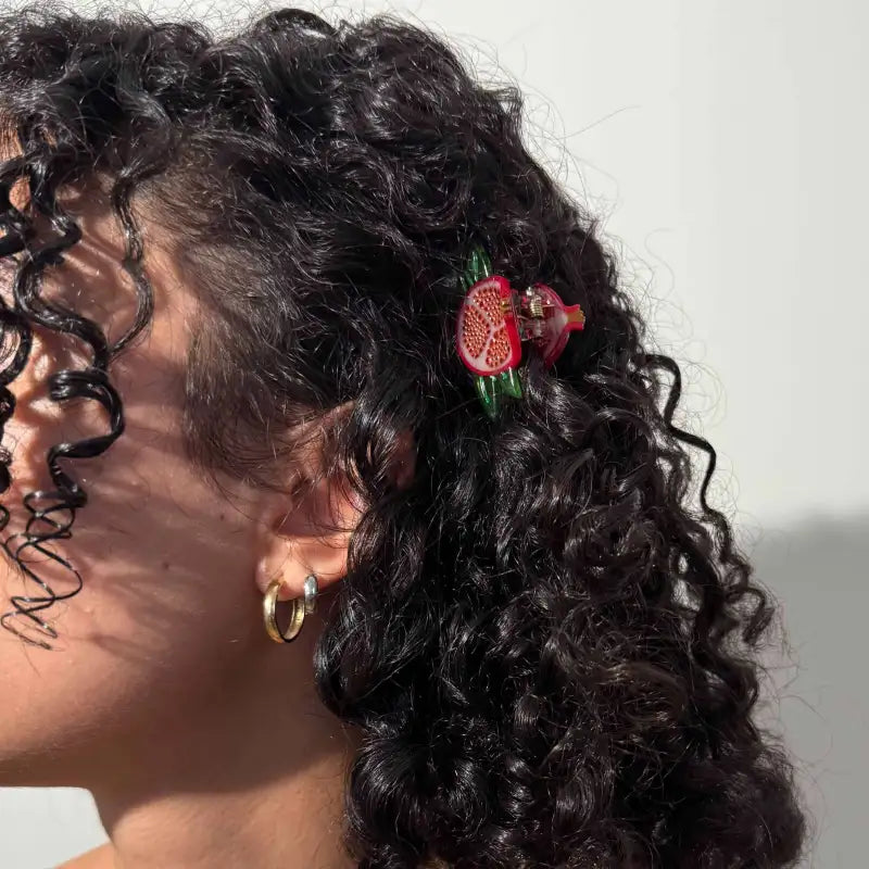 A person with dark, curly hair wearing a small red and green pomegranate-shaped hair claw clip on the side of their head.