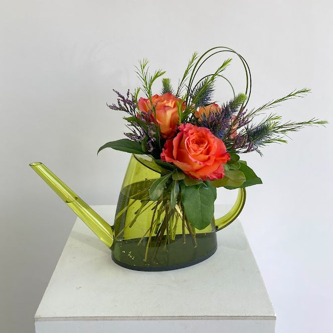 Floral arrangement in a green watering can on a white surface with a light gray background