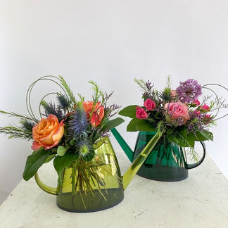 Two floral arrangements in green watering cans on a white surface.