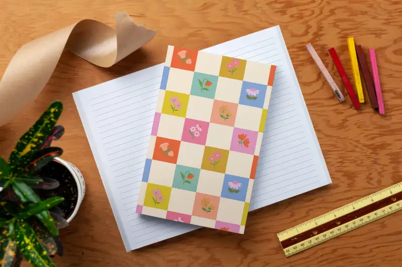 Notebook with a checkered cover featuring flowers on a wooden surface with stationery items.
