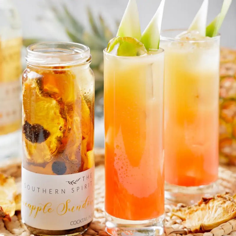 Two glasses of a colorful cocktail with a jar labeled 'The Southern Spirit Pineapple Sandia' on a textured surface.