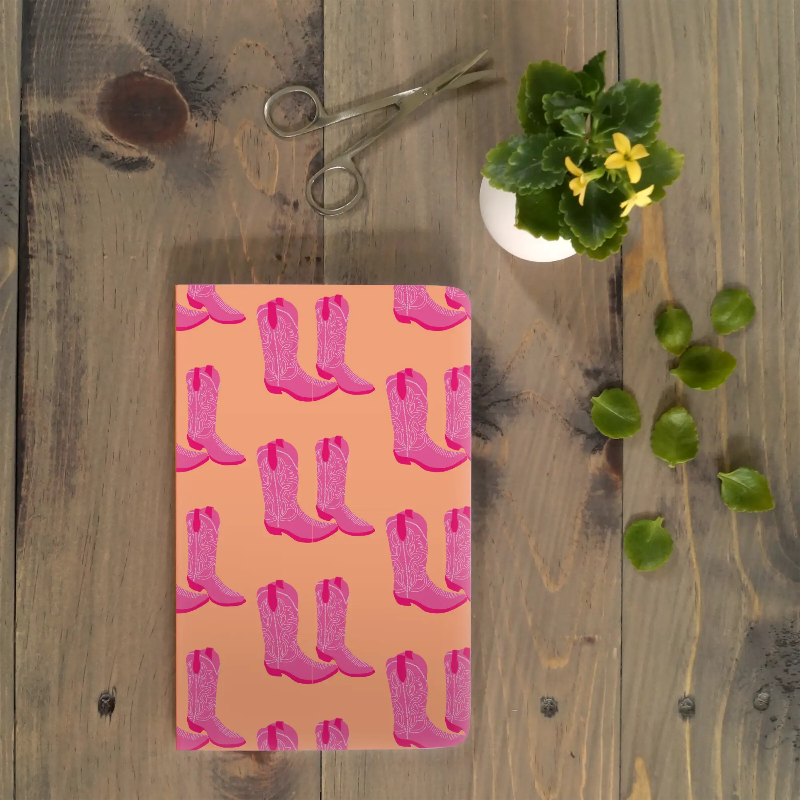 Notebook with pink cowboy boot pattern on a wooden surface with scissors and a plant.