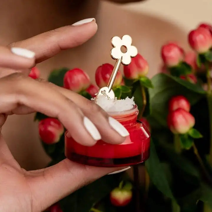 Hand holding a small red container with a white flower-shaped object, against a blurred background of red flowers.