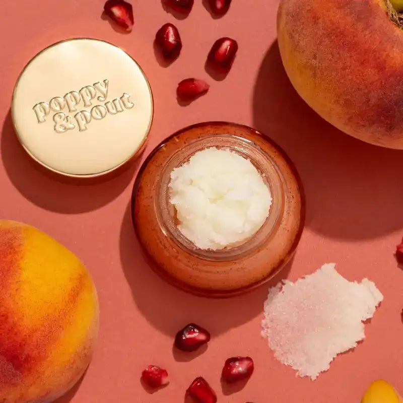 Jar of cream with 'Poppy & Pout' branding, surrounded by peaches and pomegranate seeds on a pink background.